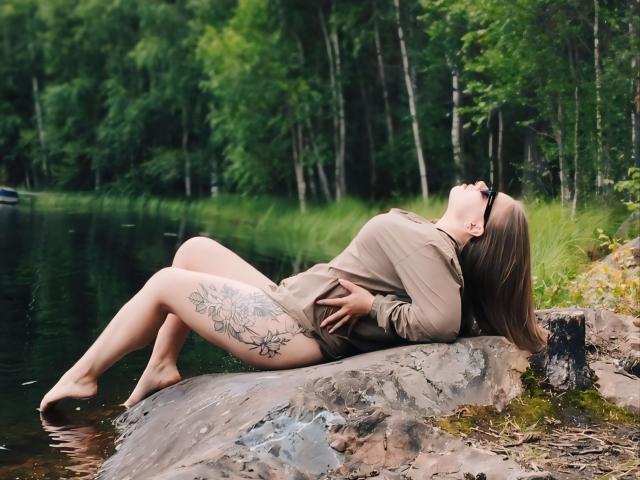 Niferttiti with tattoos on her right thigh, lying on a rock by a lake, wearing a brown long-sleeve shirt, head tilted back, legs in water, arms resting on her belly.