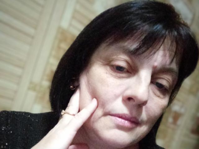 MillieSophie, middle-aged woman with black hair, slightly wrinkled face, and pink lips, touching her cheek with her right hand, wearing a black top, looking down.