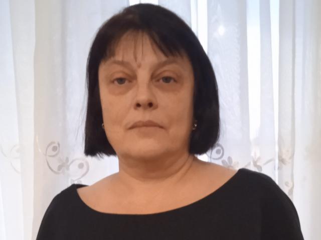 Middle-aged MillieSophie with short black bob, wearing a black top, looking straight ahead with a neutral expression, small earrings, and white curtains in the background.