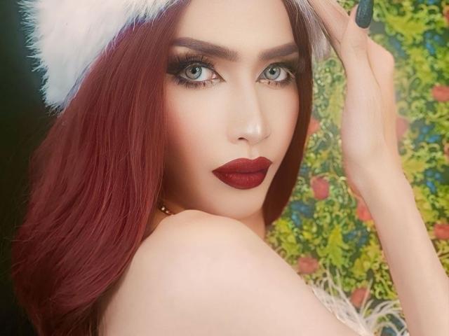 AlevaLeboria with red hair, white furry hat, dark red lipstick, blue eyes, and black nails, looking over shoulder, hand in hat. Boobs partially visible. Slight makeup, seductive expression.