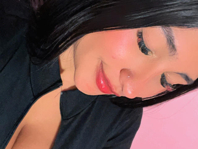 AbrilLuxurie with glossy pink lips, slight nose piercing, black hair, and closed eyes, wearing a black shirt, showing a hint of cleavage. Soft, warm skin, relaxed expression.