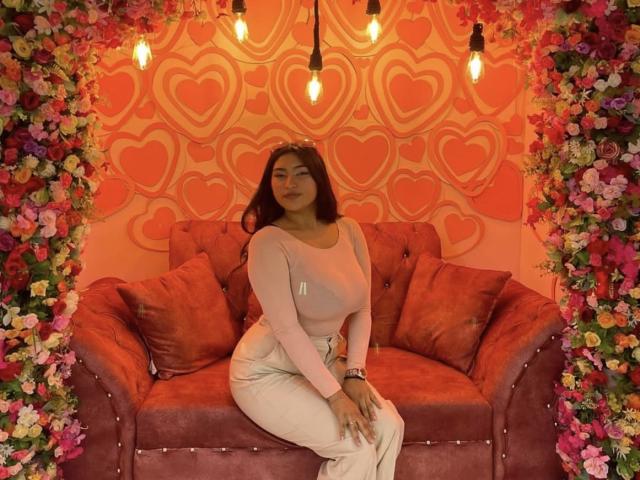 AbrilLuxurie with big ass, medium tits, long dark hair, wearing pink top and white pants, sitting on red velvet couch, surrounded by colorful flowers, under hanging lights.