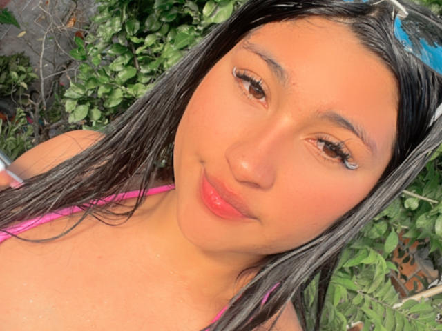 AbrilLuxurie has wet black hair, brown eyes, and pink lipstick, wearing a pink top, with a cute, slightly smug expression and her ass barely visible.