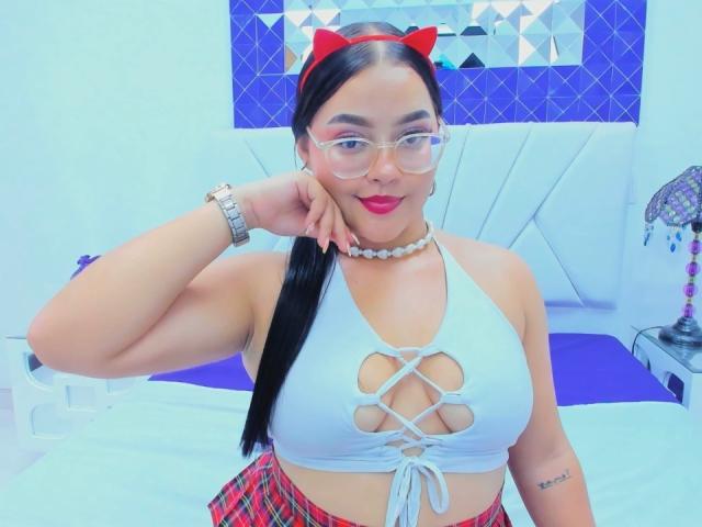 CandasLeroy with red cat ears, glasses, white lace-up top showing her big tits, red plaid skirt, and white choker, posing in a bedroom. Her black hair is in a ponytail.