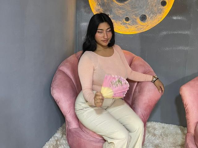 AbrilLuxurie with dark hair, pink sweater, white pants, holding pink card, sitting in pink velvet chair, smiling, thick ass, curvy, cute, wearing watch, on fluffy rug.