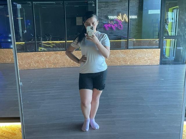 NaughtyxBunny stands in front of a mirror, taking a selfie. She's wearing a grey t-shirt, black shorts, white socks, and has her hands on her hips, showing her ass.