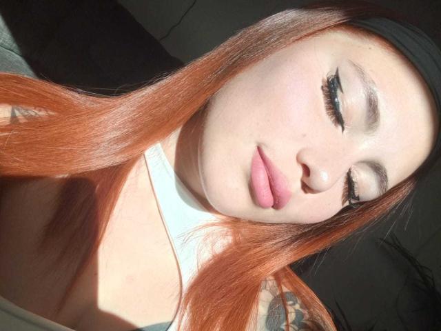 DominicTheOne has red hair, pale skin, and eyeliner with pink lips. She's wearing a black headband and white collar, with tattoos visible on her left arm.