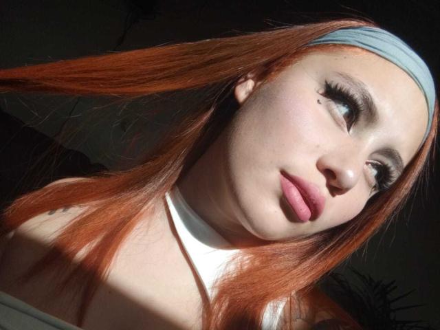 DominicTheOne has pale skin, red hair, and a grey headband. She's looking sideways with long eyelashes, pink lips, and a small mole above her upper lip. Boobs visible.