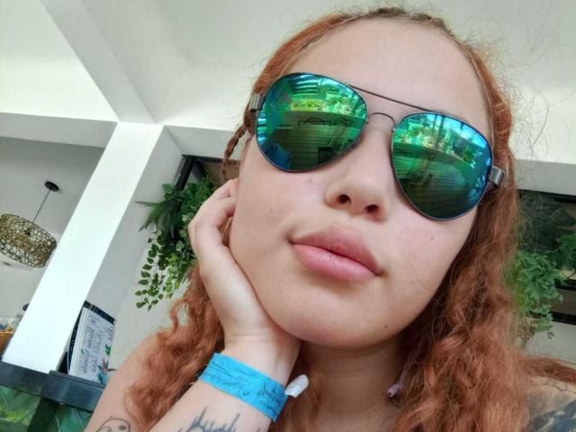 DominicTheOne has curly red hair, wearing green reflective sunglasses, and a blue wristband, resting her head on her hand with a subtle smile. Her pink lips are slightly parted.
