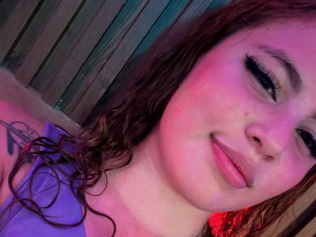 DominicTheOne has pink-tinted skin, wavy brown hair, black eyeliner, and a small tattoo on the upper arm. She's smiling with pink lips, wearing a purple top.