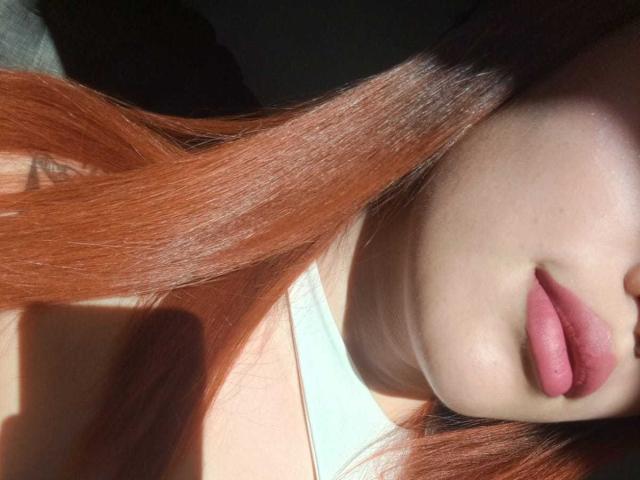 DominicTheOne's face with light skin, pink lips slightly parted, and long red hair, sunlit, wearing a white top, showing a small part of her upper chest.