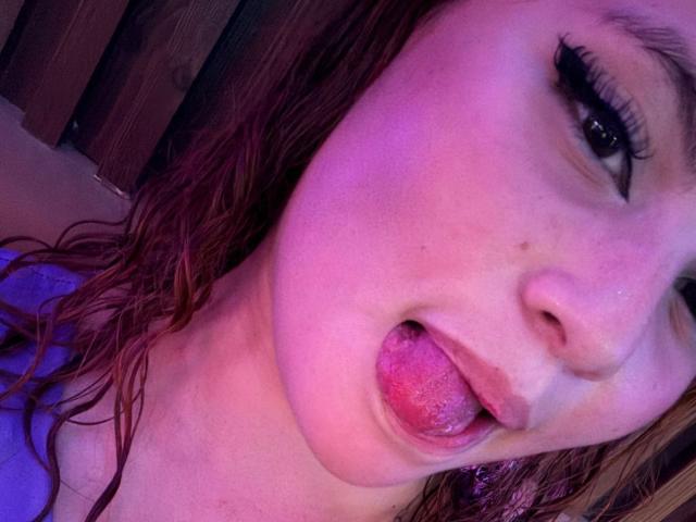 DominicTheOne's face with pink-tinged skin, wet red hair, and tongue sticking out. Black eyeliner, slightly open mouth, visible tongue. Background wood and purple light.
