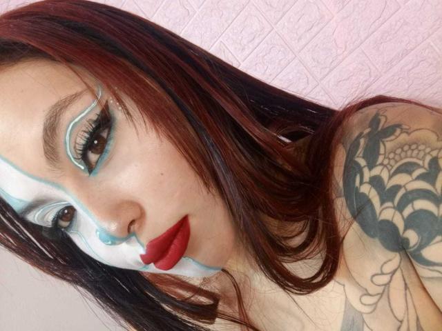 DominicTheOne has red lipstick, blue and white face paint, dark red hair, and a tattoo on their shoulder. Their tits are partially visible, and they have a nose ring.