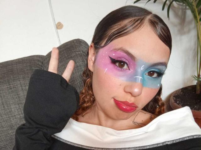 DominicTheOne has colorful face makeup with purple on the left, blue on the right, and white dots, brown curly hair, red lipstick, and is giving a peace sign.