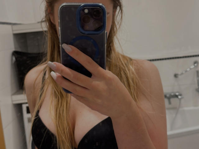 SunnyBeatrice with long blonde hair, wearing a black strapless top, showing her boobs in a mirror selfie, holding a blue phone with a circular grip.