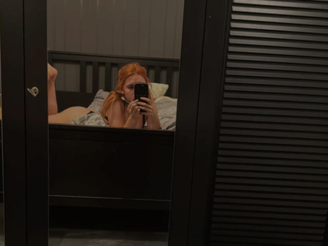 SunnyBeatrice with red hair lies on a bed, taking a mirror selfie, showing her bare ass and a glimpse of her tits, holding a black phone.