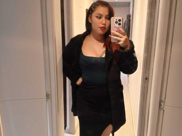 DominicTheOne with red hair, wearing a black coat, green top, and black skirt, showing a thigh cutout, taking a mirror selfie with a phone, showing her tits.