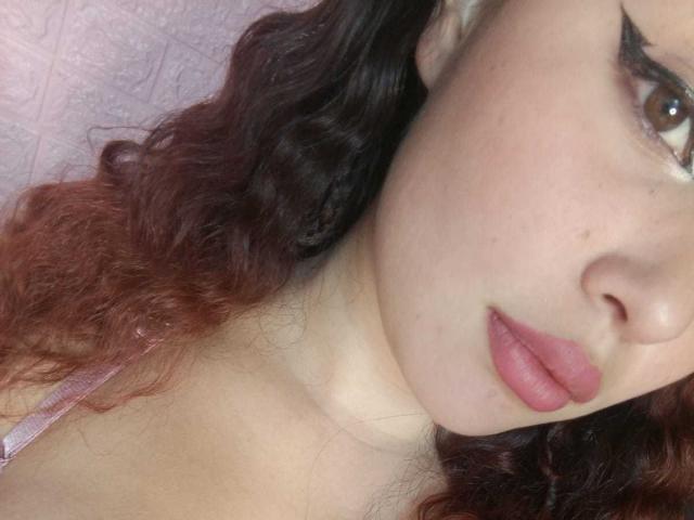 DominicTheOne's face, showing wavy red-brown hair, pale skin, thick eyebrows, pink lipstick, and a slightly open mouth. Boobs partially visible, wearing a pink strap.