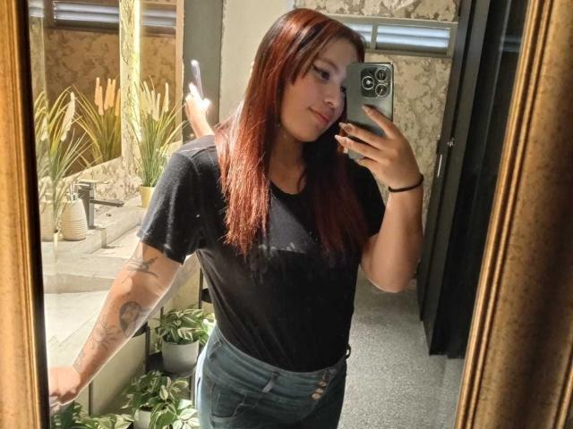 DominicTheOne with red hair, black tee, and blue jeans, showing tattoo on arm, taking mirror selfie, holding phone, wearing black bracelet, in a well-lit room.