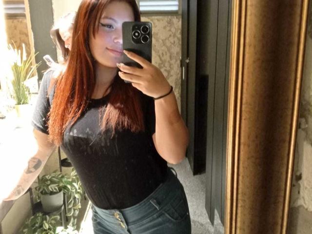 DominicTheOne with red hair, black t-shirt, and green jeans, taking a mirror selfie, showing tattoos on her right arm, holding a black phone, smiling slightly.