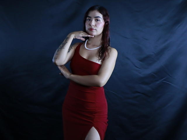 Eileen Swift, con un vestito rosso, una collana bianca e tatuaggi, in piedi con una mano sul mento, che guarda la fotocamera, su uno sfondo scuro, mostrando il suo sedere con una spaccatura alta fino alla coscia..