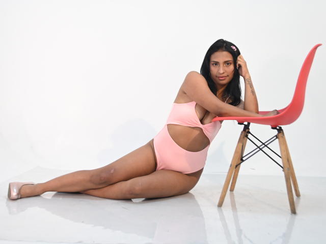 ValentinaVera with brown skin, black hair, pink one-piece, sitting on floor, leaning on red chair, showing ass, legs stretched out, looking at camera, wearing pink heels.