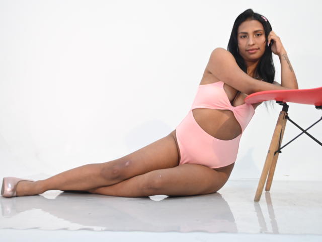 ValentinaVera with tan skin, long black hair, pink cut-out swimsuit, showing her tits and ass, sitting on floor, legs stretched, leaning on red chair, wearing pink heels.