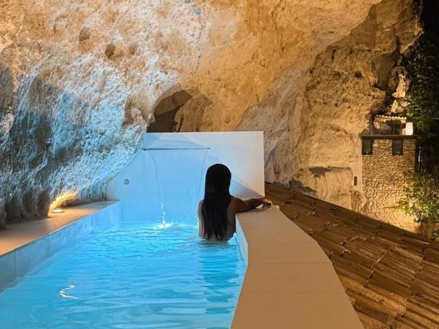 JennerSex with long black hair, back to the camera, in a small blue-tiled indoor pool, standing up, showing her ass above the water, against a rocky cave wall.