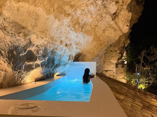 JennerSex has a dark butt in a blue-tinted pool, sitting with back to camera, in a rocky cave. Cave walls are textured, with small lights on left.