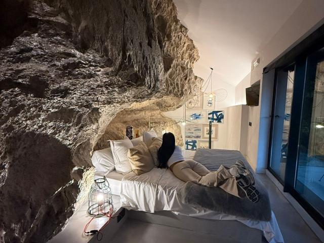 JennerSex è sdraiata su un letto bianco con cuscini, con i glutei rivolti verso l'alto, indossa abiti bianchi e neri, in una stanza con una parete rocciosa e una lanterna sul pavimento..
