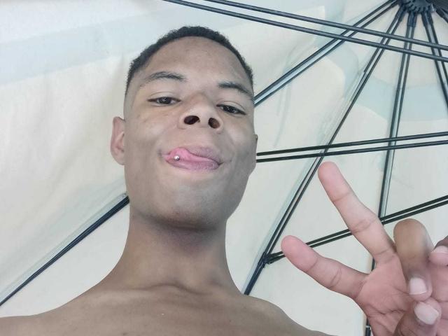 MasterXx, shirtless, dark skin, short hair, sticking out tongue, small pink lip piercing, making a peace sign, umbrella with black metal frame in the background.