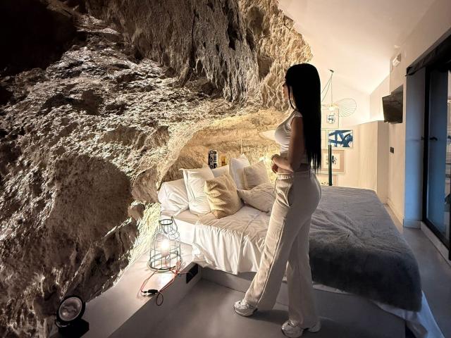 JennerSex est debout, les fesses face à la caméra, vêtue d'un pantalon blanc et d'un débardeur, dans une pièce avec un mur de rochers, un lit avec des oreillers et une petite table avec des objets. Elle a de longs cheveux noirs..