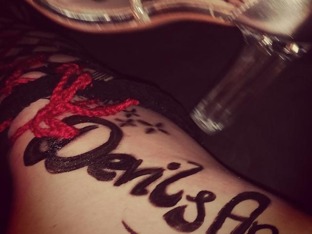 O traseiro tatuado de DevilsAngel tem uma escrita preta com um detalhe em vermelho que se assemelha a uma corda, ao lado de um copo, tudo sob uma luz suave e quente. O estilo da tatuagem é ousado e cursivo..