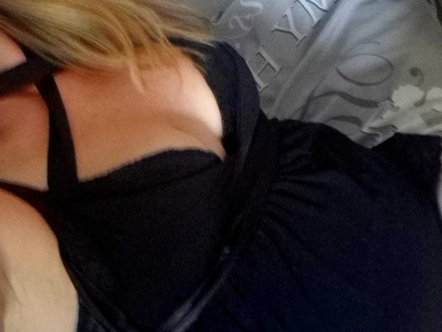 El vestido negro con tirantes de DevilsAngel muestra sus grandes senos, con un escote bajo y un corte ligeramente revelador, mientras está acostada y su cabello rubio se ve parcialmente..