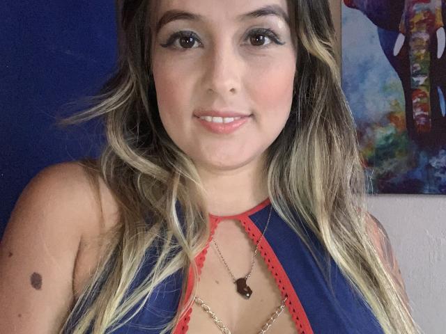 SaritaCaramelo with long blonde and brown wavy hair, wearing a blue and red halter top, smiling, and showing a small black mole on her right shoulder.
