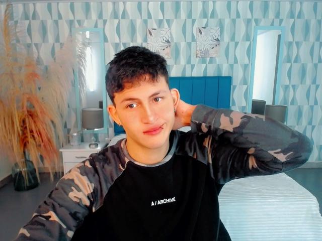 AxelSavage, un joven con cabello corto y oscuro, que lleva una camisa de manga larga con estampado camuflado en negro y gris, tiene una mano detrás de la cabeza y mira a la cámara con un ligero puchero..