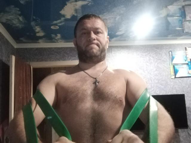 Shirtless AbubakarH with a beard, green suspenders, chest hair, and a necklace, standing in a room, looking down at the camera with a serious expression.