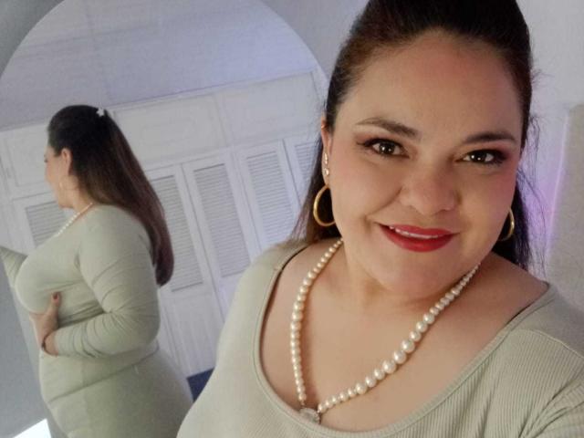 NatashaBlade with brown hair in a ponytail, wearing a green long-sleeve top, pearl necklace, and red lipstick, smiling at the camera, reflected in a mirror showing her ass.