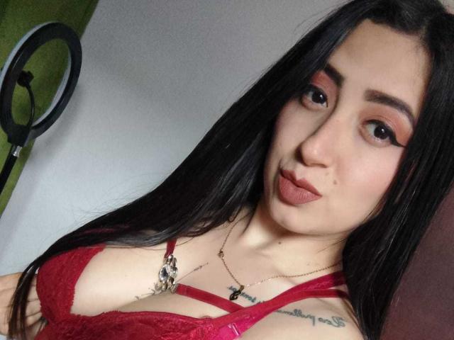 JuliethCruz with long black hair, red lace bra, pouty lips, and tattoos, wearing a necklace, looking at the camera, ring light on the left. Boobs are visible.