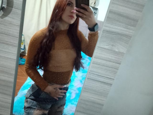 SofiaFountaine with long red hair, wearing a sheer brown top, showing her tits, holding a phone, wearing camo pants, in front of a mirror.