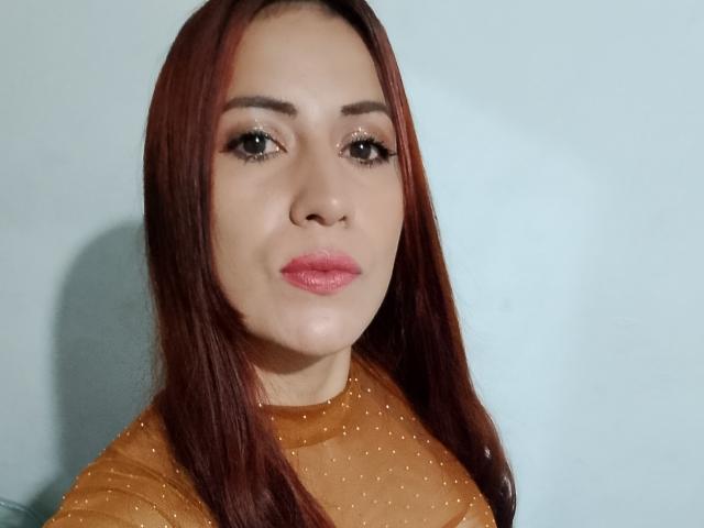SofiaFountaine with red hair, wearing a sheer brown top, has pink lipstick and dark eyeliner, looking at the camera with a neutral expression, no visible boobs or pussy.