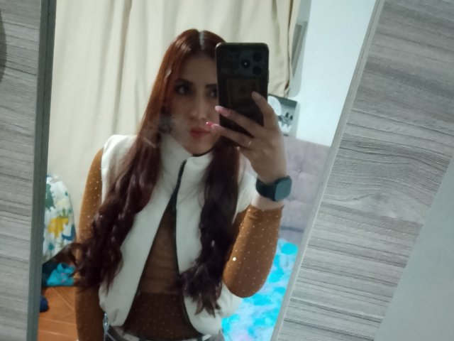 SofiaFountaine with long red hair, white scarf, brown top, holding phone in mirror selfie, wearing blue watch, pink lipstick, reflective mirror, wooden texture on right.
