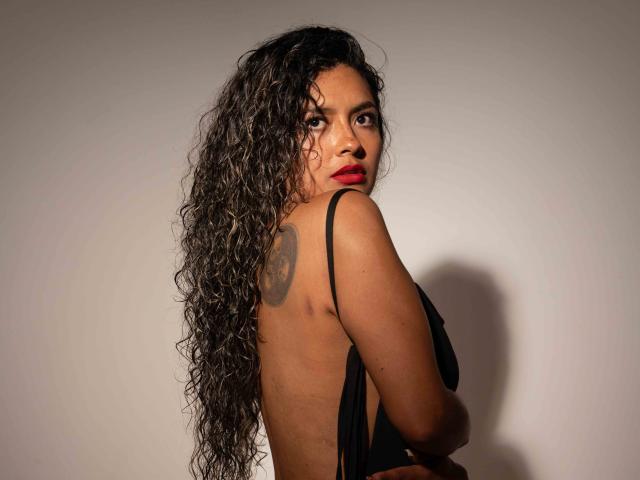 JedaZore mit langen, lockigen, schwarzen Haaren, rotem Lippenstift und einem schwarzen Top ohne Rücken, das ein Tattoo auf ihrem oberen Rücken zeigt, blickt über ihre Schulter. Ihre braune Haut wird hervorgehoben..