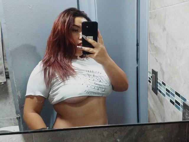 LilithMoonryn with red hair, lifting her white shirt to expose her large tits, taking a mirror selfie in a bathroom, holding a black phone.