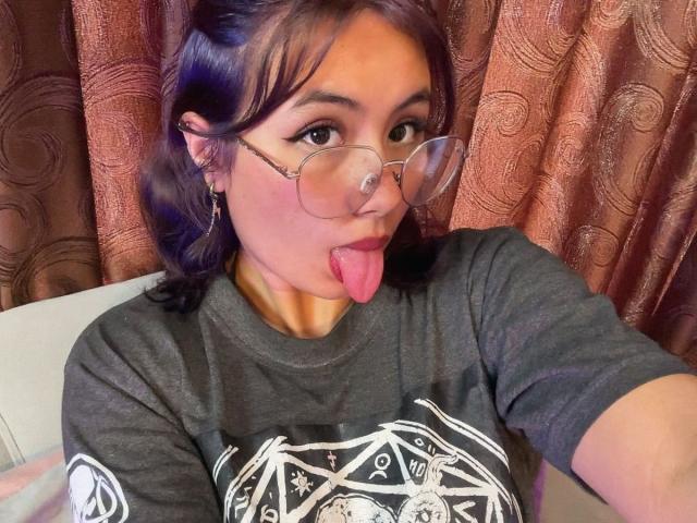 NovaAllen with purple hair, glasses, and tongue out, wearing a gray shirt with white print, has her tongue sticking out, showing her face and upper body.