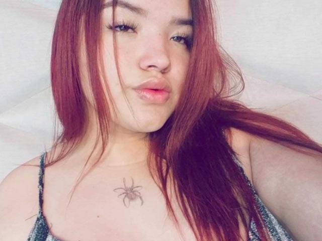 LilithMoonryn with long red hair, pouty lips, and a spider tattoo on her chest, wearing a sparkly top, looking at the camera. Her tits are slightly visible.