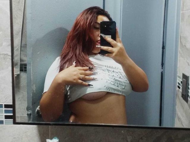 LilithMoonryn with red hair, lifting her shirt to show her tits in a bathroom mirror selfie, holding a black phone, wearing a gray shirt, looking at the camera.