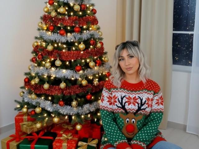 EmilyValk with blonde hair, wearing a red and green Reindeer Christmas sweater, sitting next to a decorated Christmas tree with red, gold, and silver ornaments.