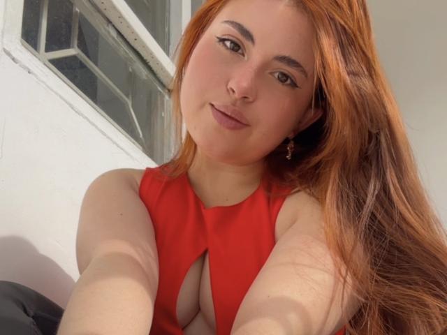 VicluX mit langen roten Haaren, trägt ein rotes Kleid mit einem Ausschnitt, lächelt in die Kamera und hält ihr Handy hoch, um ein Selfie zu machen. Ihre Brüste sind teilweise sichtbar..