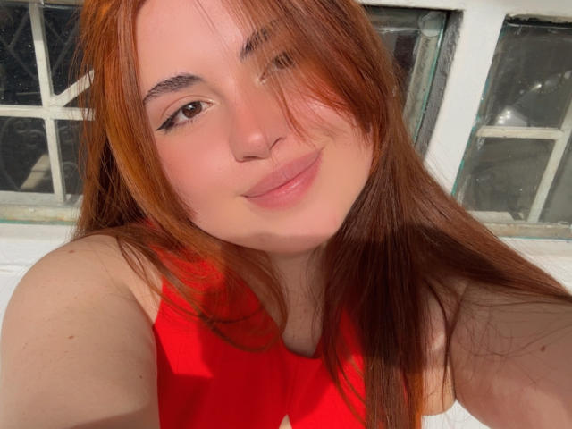 VicluX with red hair, smiling, wearing a red top, has light skin, brown eyes, and a small nose. Her hair's slightly messy, and she's looking at the camera.